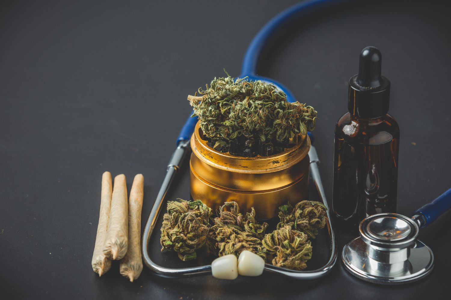 Three joints, a grinder with flower, a tincture bottle and a stethoscope sit on a dark surface.