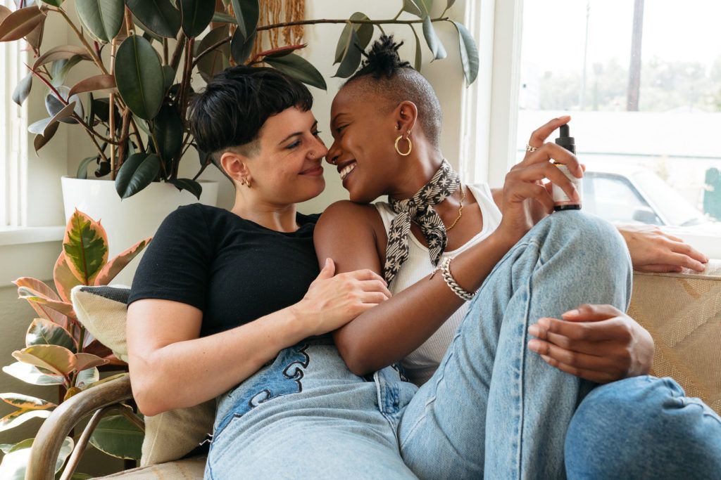 Two women embrace intimately on a couch in a bright room with plants. They smile, touching noses. One holds a spray bottle.
