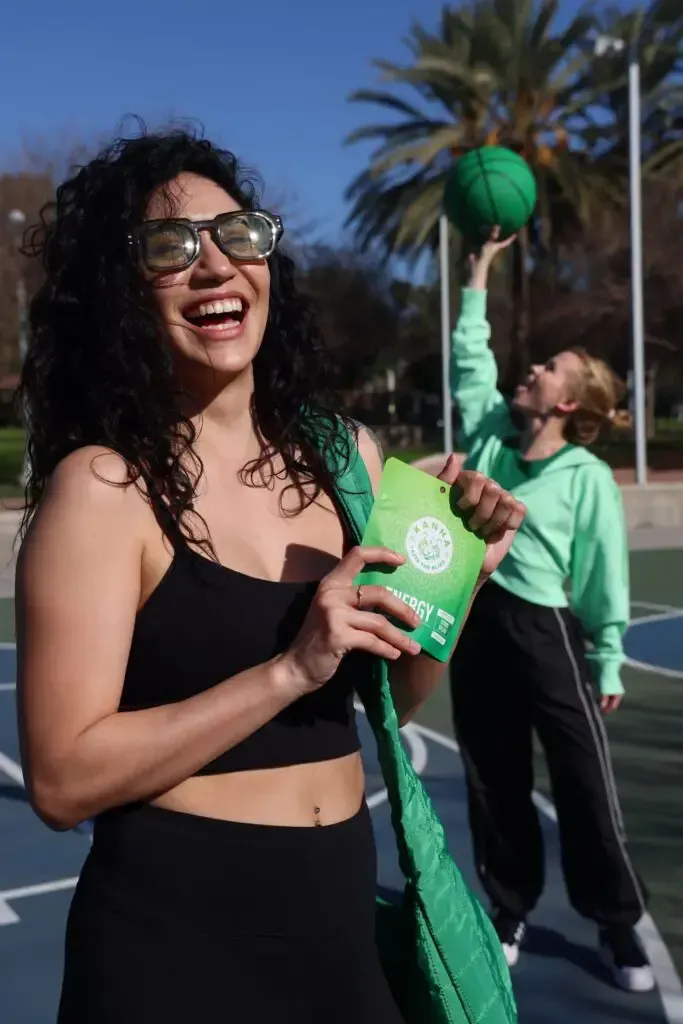 Two women on an outdoor basketball court. One laughs, holding a green bag; the other throws a green basketball.