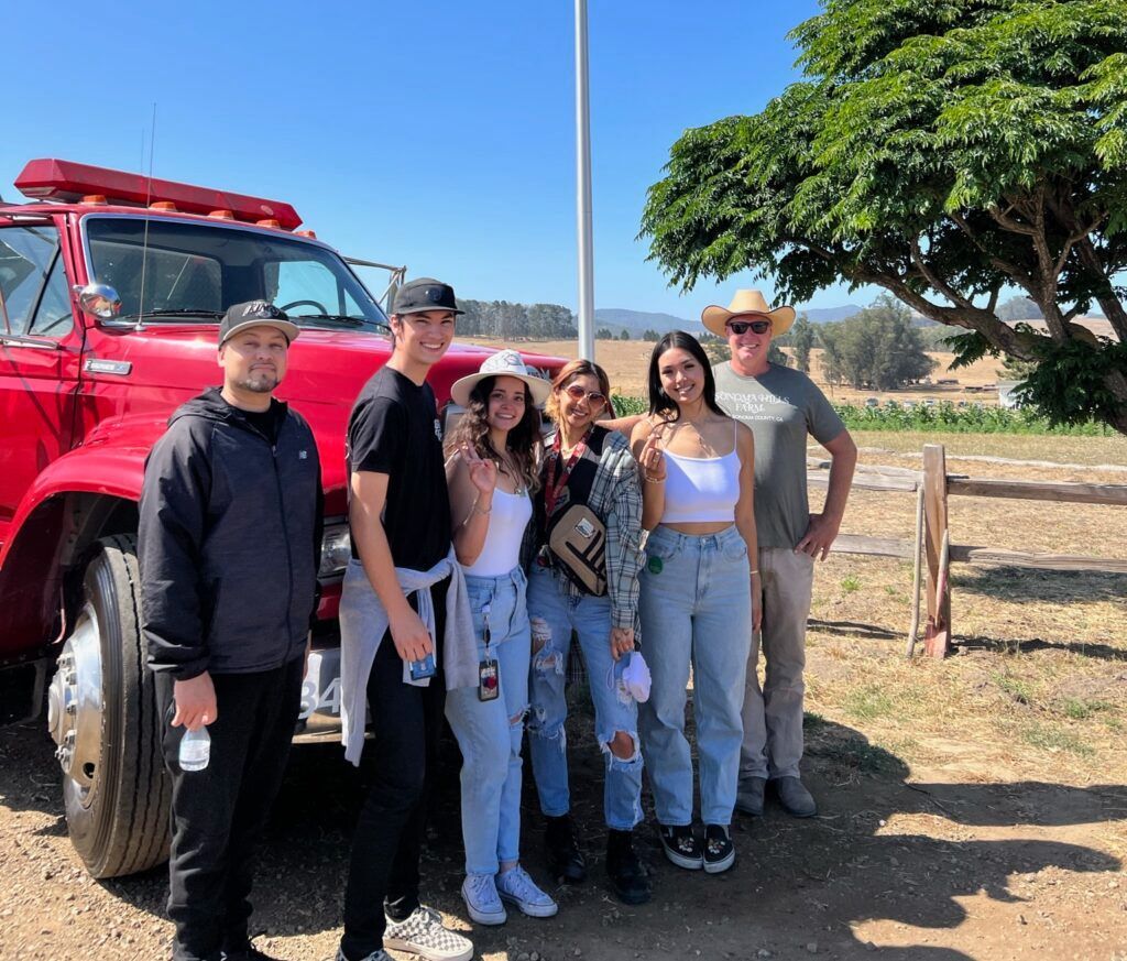 A group of seven people pose near a red fire truck on a sunny day. They stand in a field at Sonoma Hills Farm.