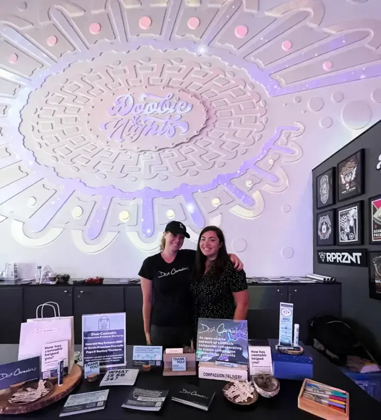 Two women stand behind a table in a shop, smiling and handing out Dear Cannabis branded bags to patients.