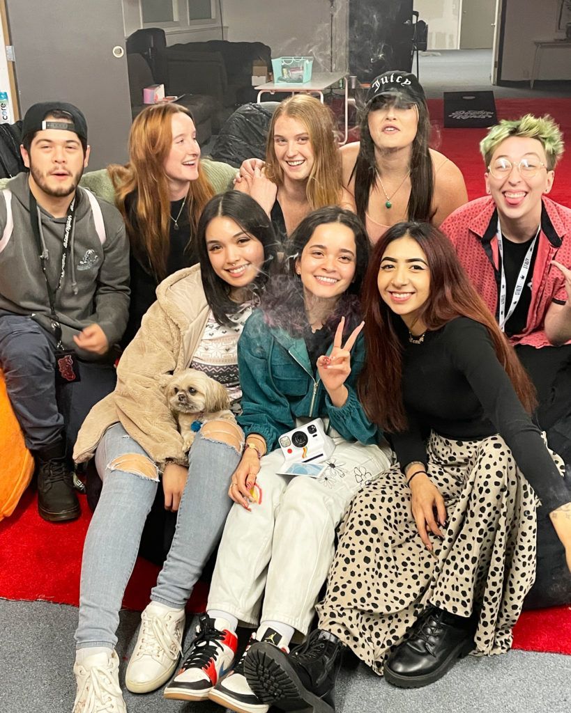 A group of nine young people smile and pose for a photo indoors on red carpet. Diverse expressions, casual clothes.
