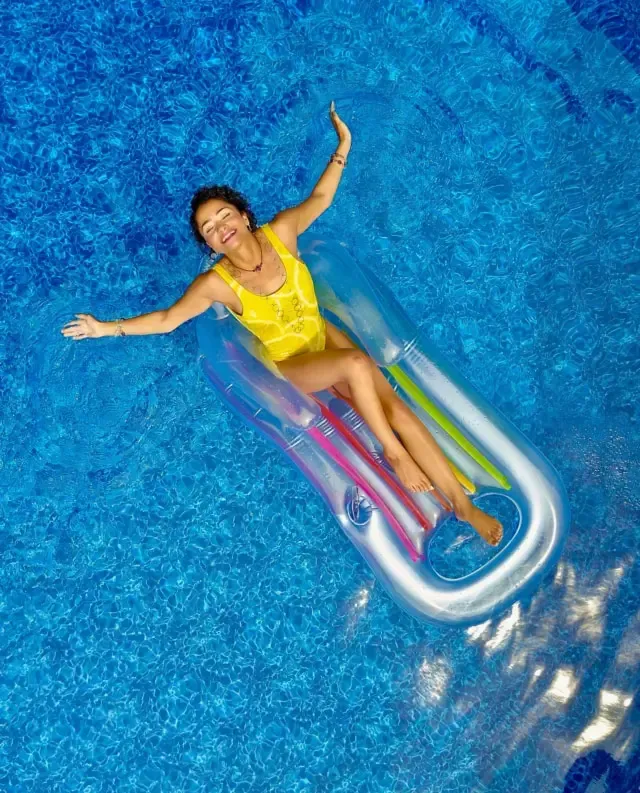 Woman in yellow swimsuit floats on a colorful raft in a bright blue pool, arms outstretched, smiling.