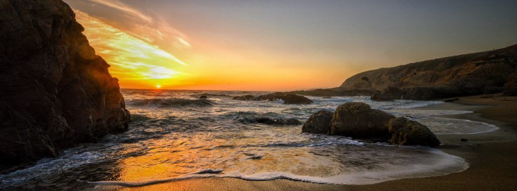 Sunset over a rocky beach. The sky is ablaze with orange and yellow hues, reflecting on the water as waves lap the shore.