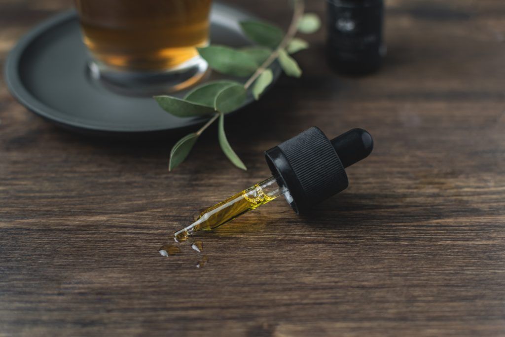 A dropper of CBD oil on a wooden surface with a glass of tea and eucalyptus sprig.