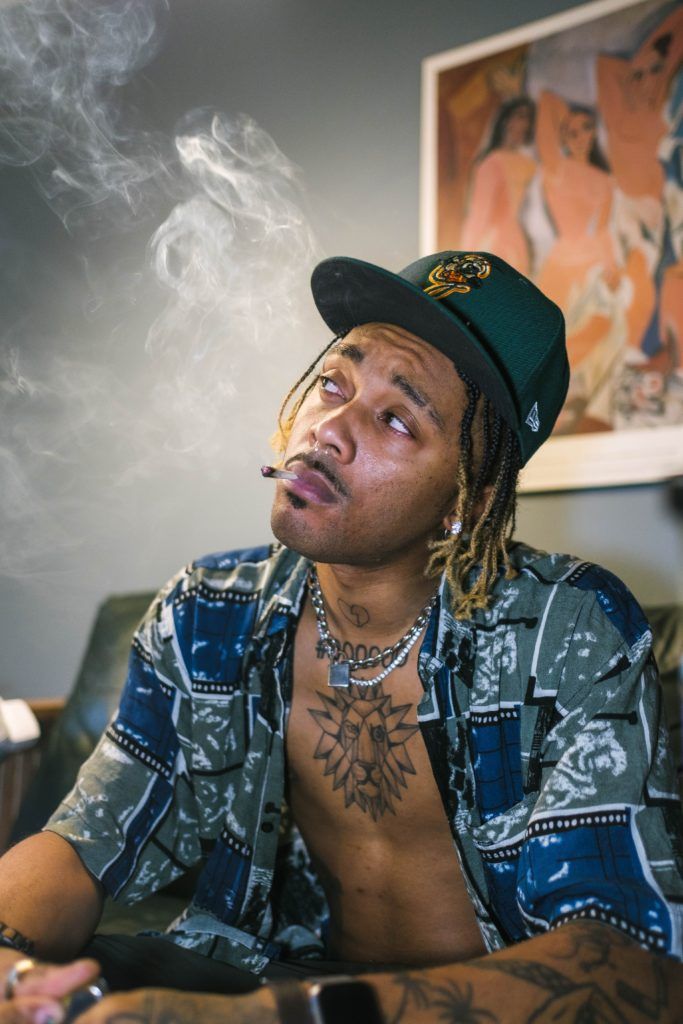 Man with dreadlocks, smoking, wearing a patterned shirt and a cap, sitting indoors, looking up. Smoke visible.