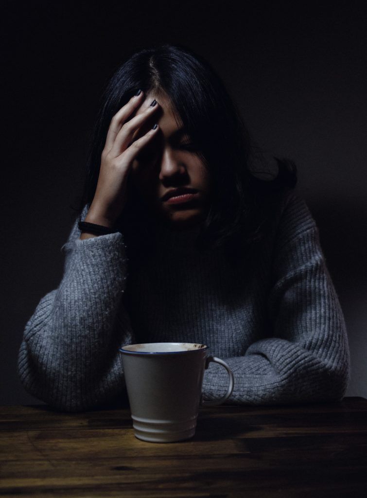Woman with hand on forehead, looking down, sitting at a table with a cup. Dark lighting, somber mood.