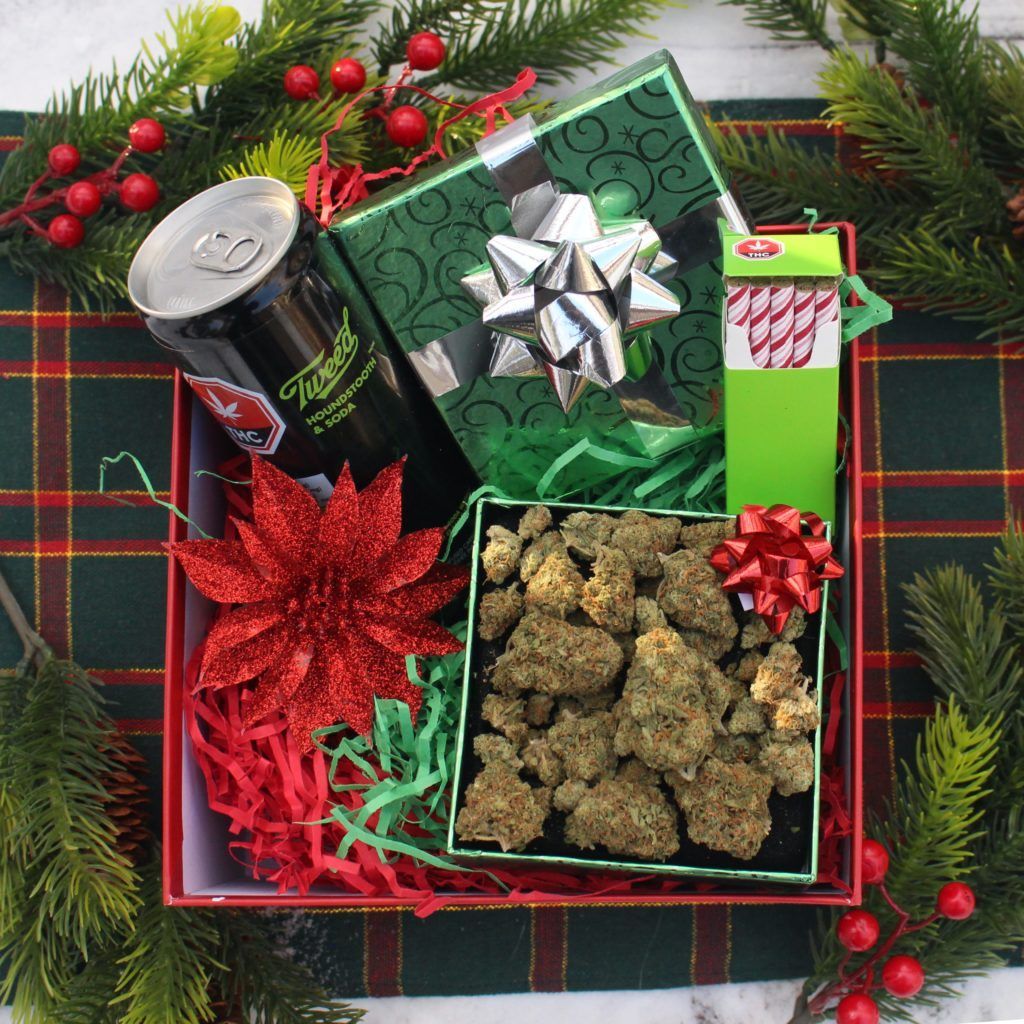 Christmas gift box with cannabis buds, drinks, and festive decorations including a poinsettia flower and wrapped gifts.