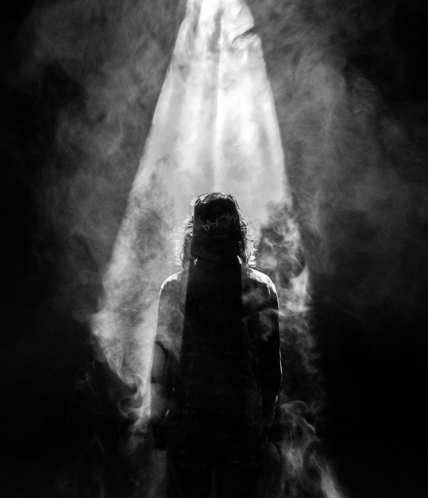 A person stands in a beam of light surrounded by smoke, silhouetted against a dark background.