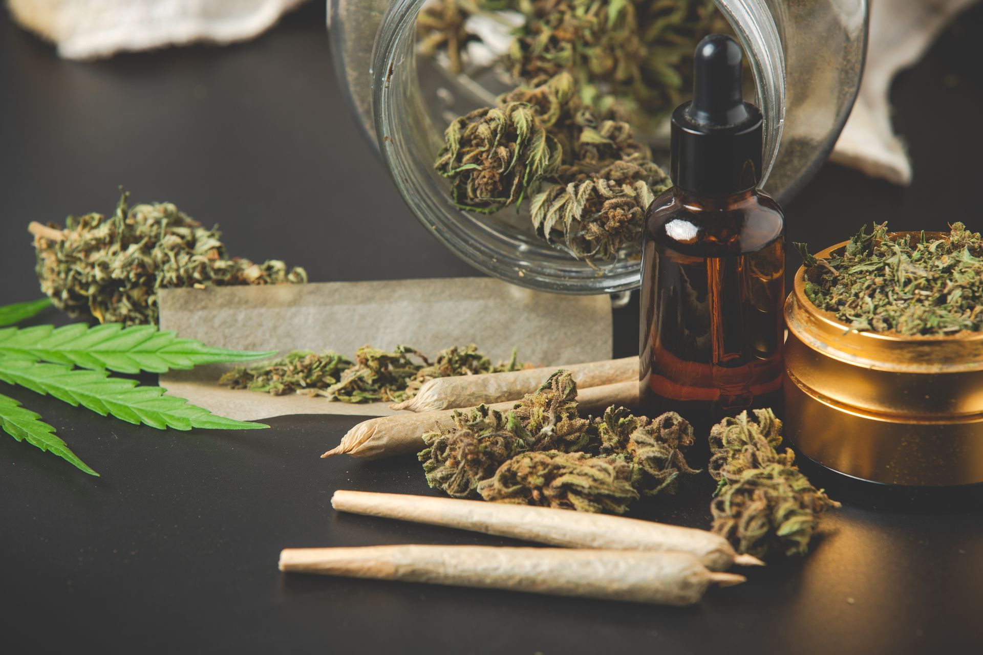 A jar of cannabis flowers spills onto a dark surface where there is a grinder, rolled joints, a tincture jar and grinder.