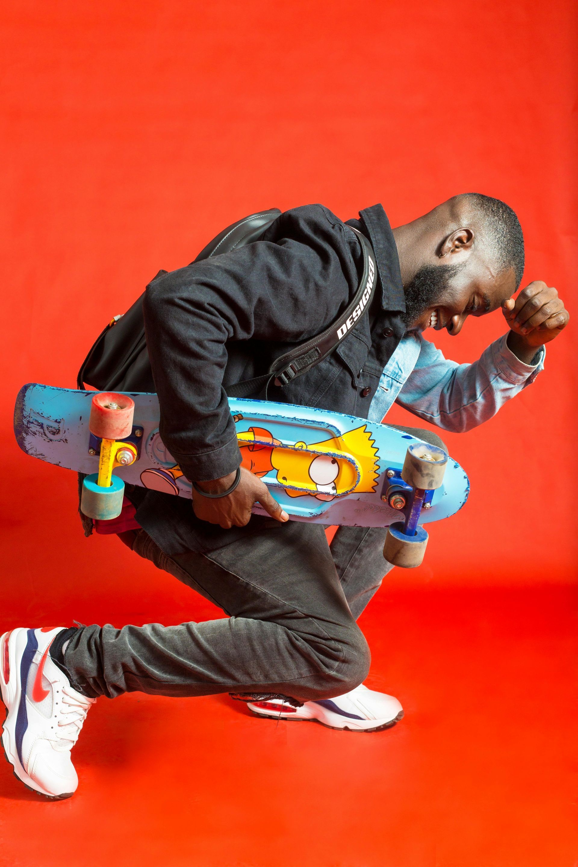Man in blue and black holds a skateboard with a Simpsons design against a red background, with an arm raised.