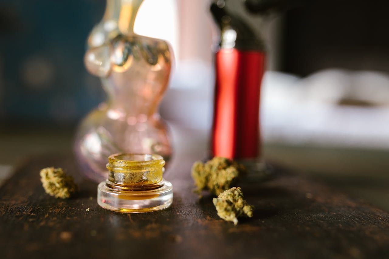 A jar of concentrates sits on a dark table with some nugs, a torch and a glass rig in the background.
