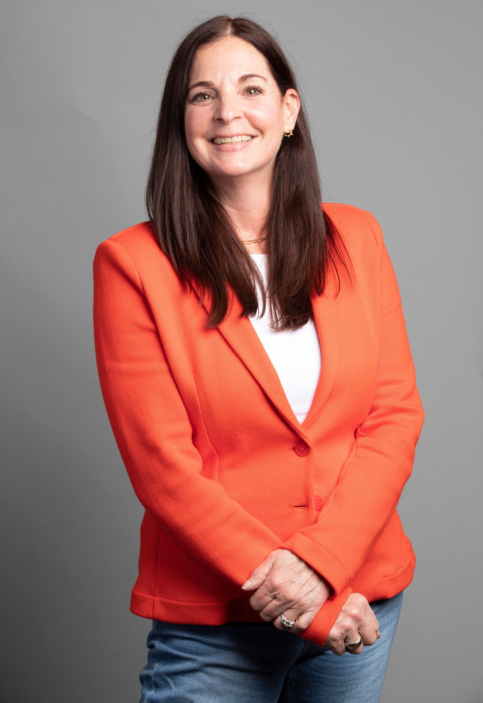 Woman in a bright orange blazer smiling against a gray studio background.