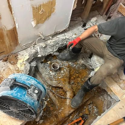 Worker in a damaged floor opening, using a shovel amid debris beside a blue fan.