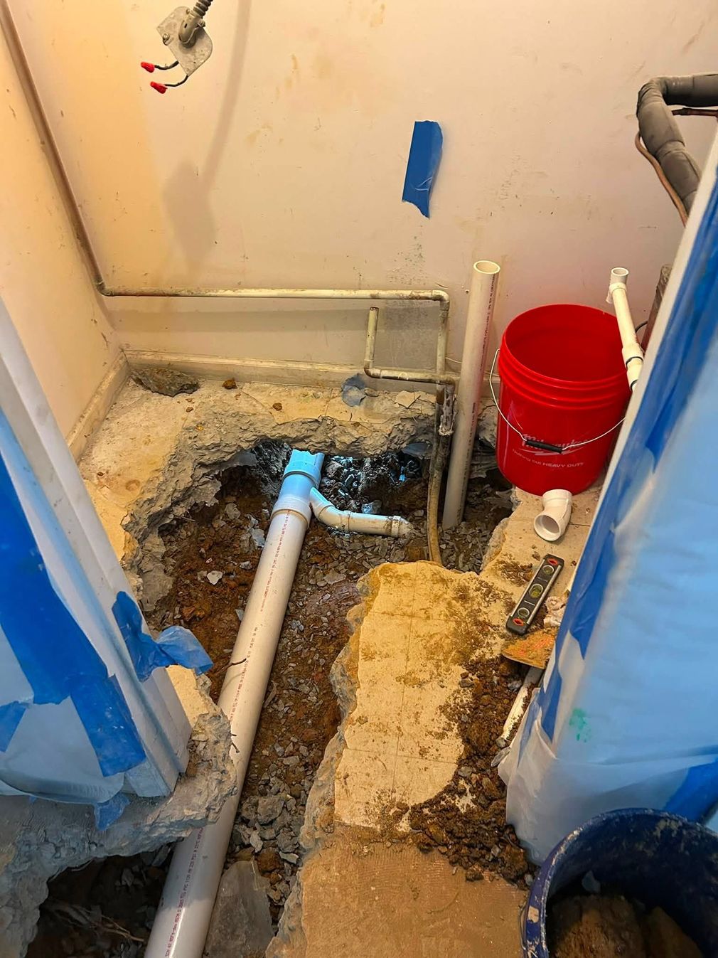 Damaged bathroom floor with exposed pipes, debris, and a red bucket, bordered with blue tape