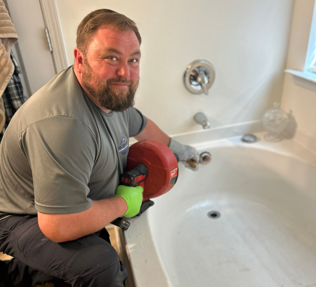 Man kneeling beside a white bathtub, holding a red object and smiling toward the camera.