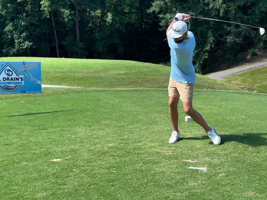 Golfer mid-swing on a sunny course, wearing a blue shirt and khaki shorts, with a blue tournament sign nearby