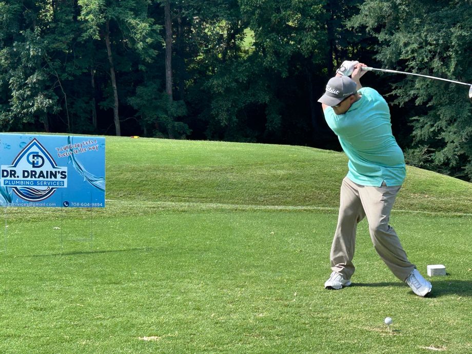 Golfer in teal shirt swings a club on a grassy course beside a Dr. Drain banner