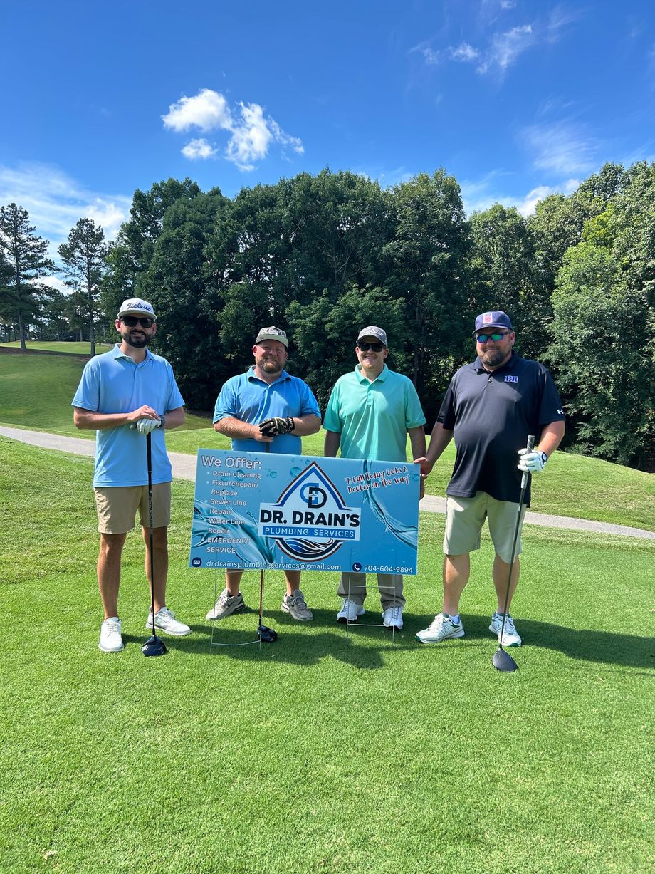 Four people holding a blue event banner on a sunny golf course, with trees and a pond behind them.