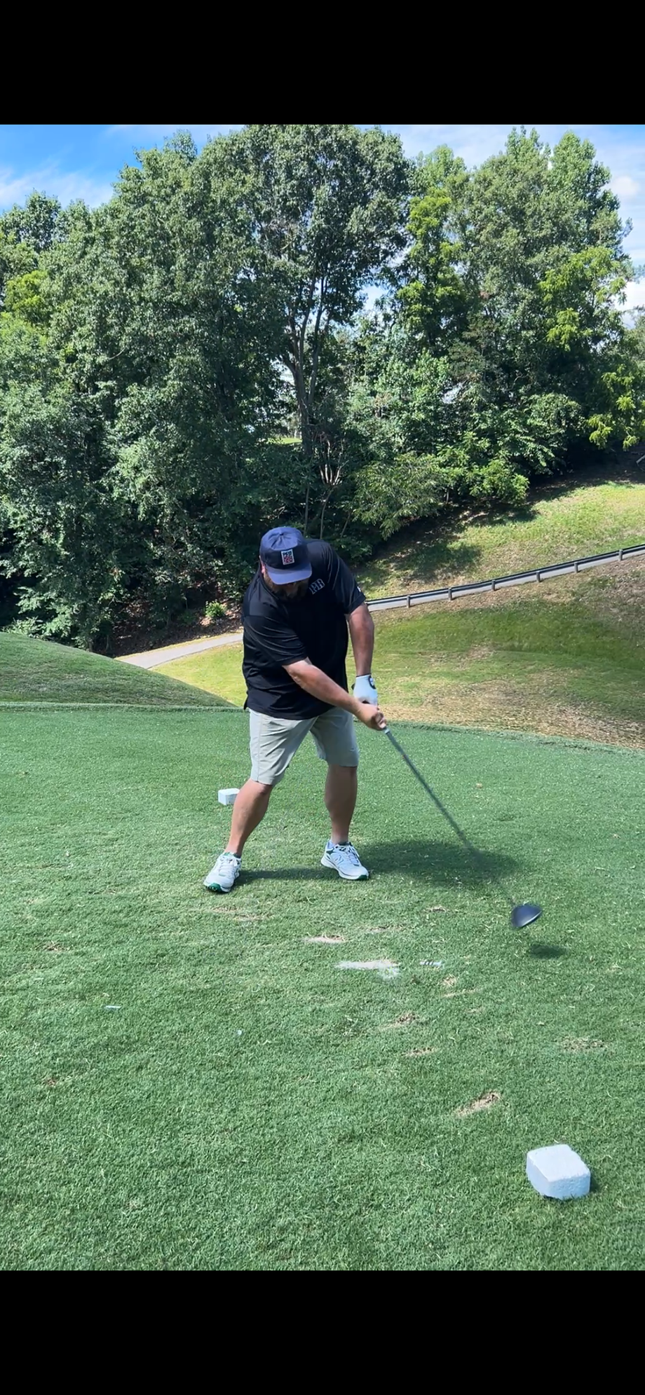 Golfer in black shirt and cap swinging a club on a grassy course with trees in the background