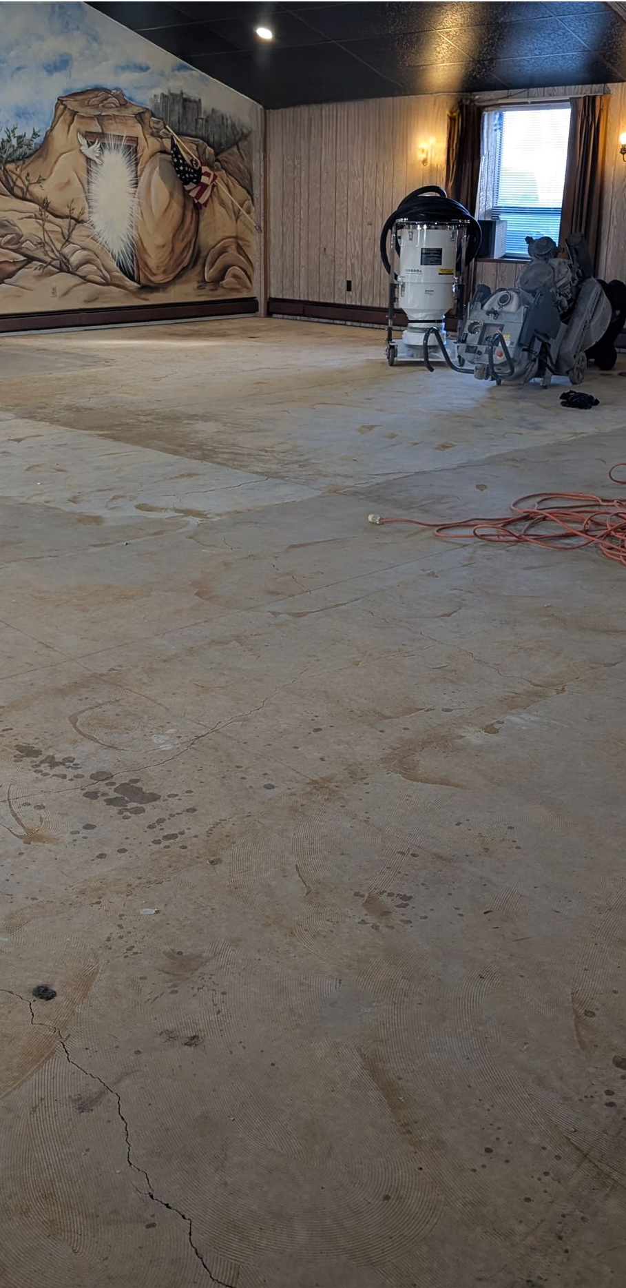 Indoor hallway with a dusty concrete floor, murals on the walls, and cleaning equipment near a doorway.