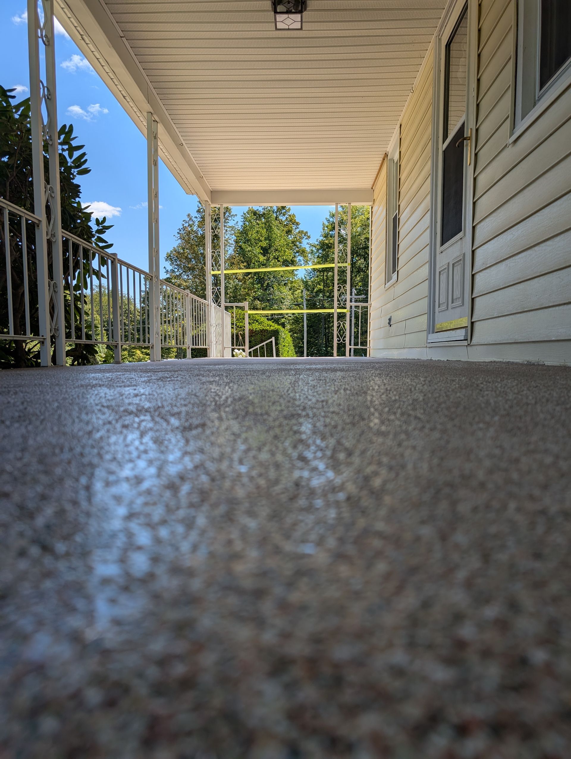 Low-angle view of a covered front porch with white siding, railing, and trees beyond.