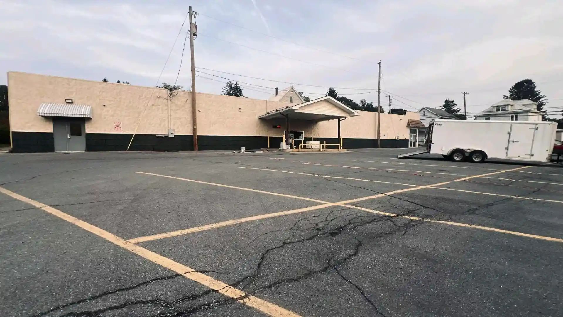 Empty cracked parking lot beside a beige building with a white trailer and utility poles under a cloudy sky