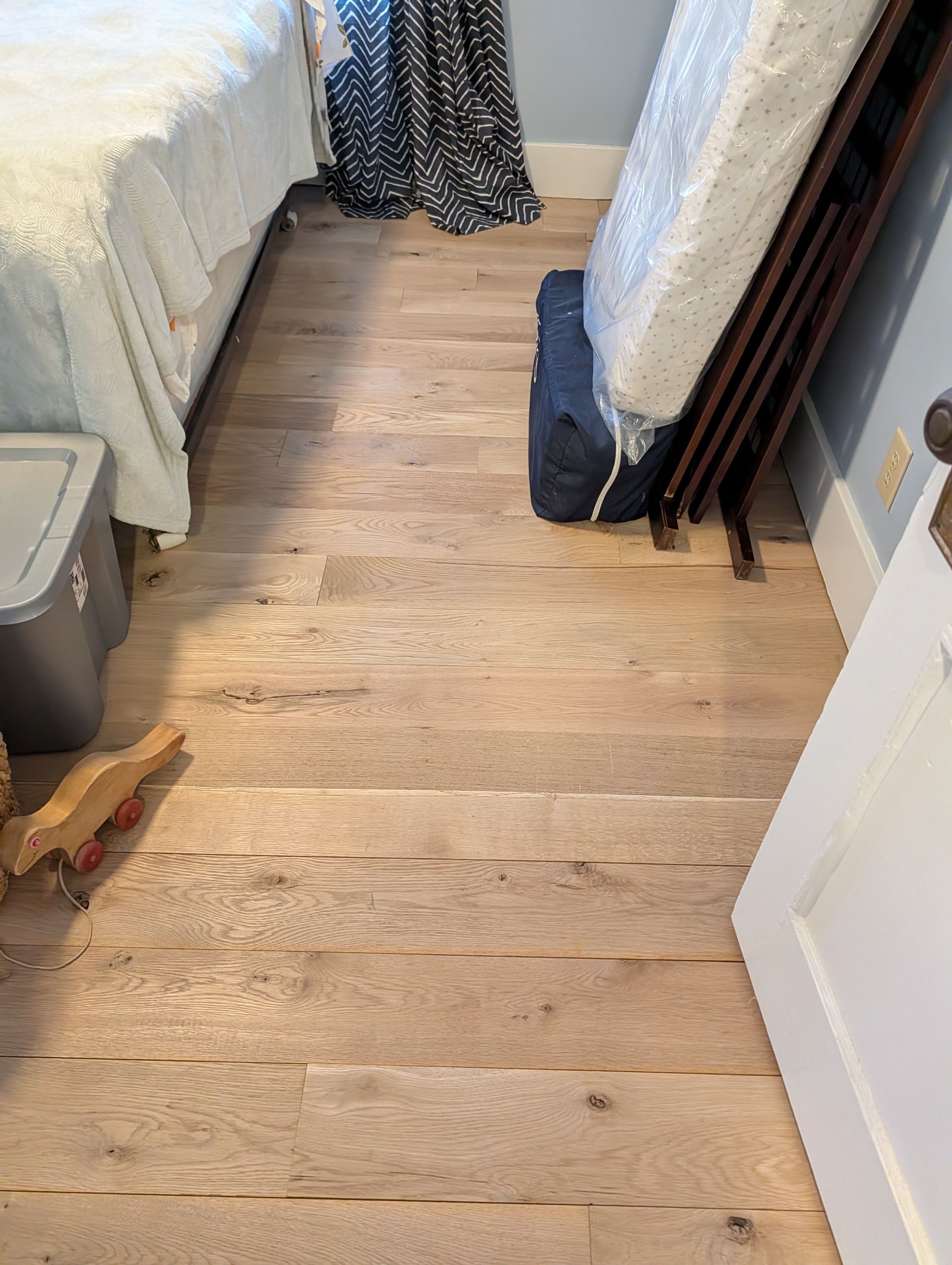Bedroom with light wood floor, white bedding, a dress hanging, and stacked long boards by the wall.