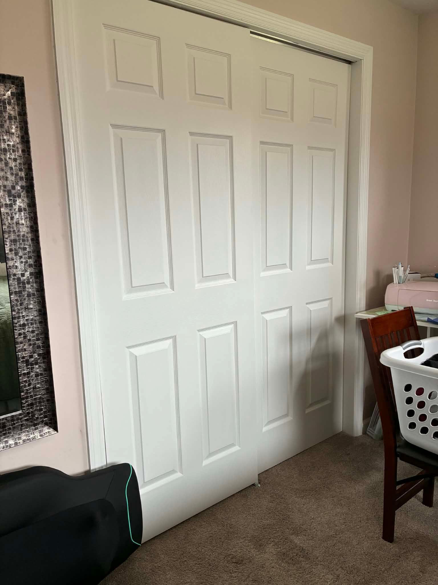 White double closet doors in a carpeted bedroom or hallway beside a window and chair.