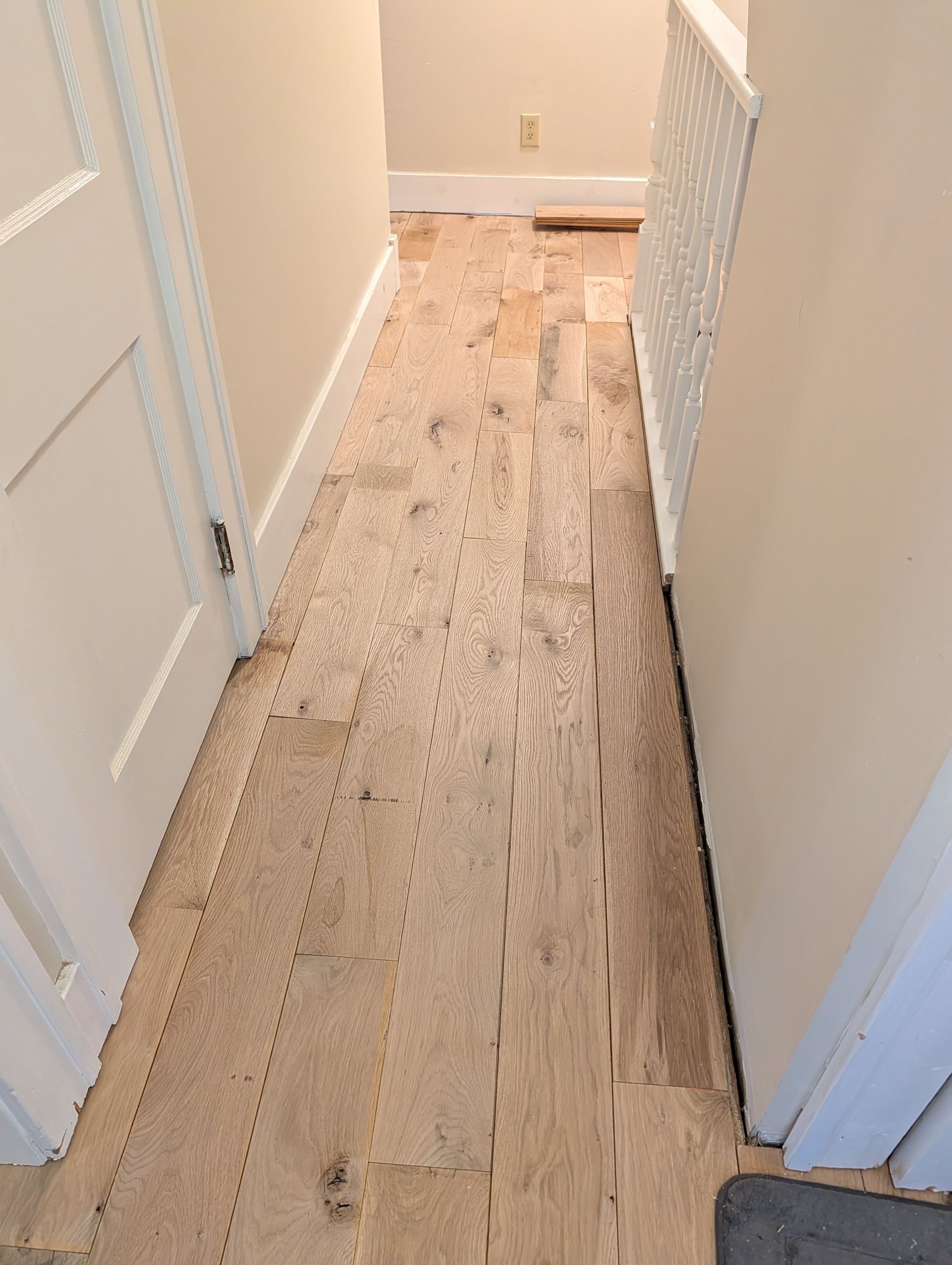 Narrow hallway with a worn, damaged wooden floor and white walls, viewed from one end.