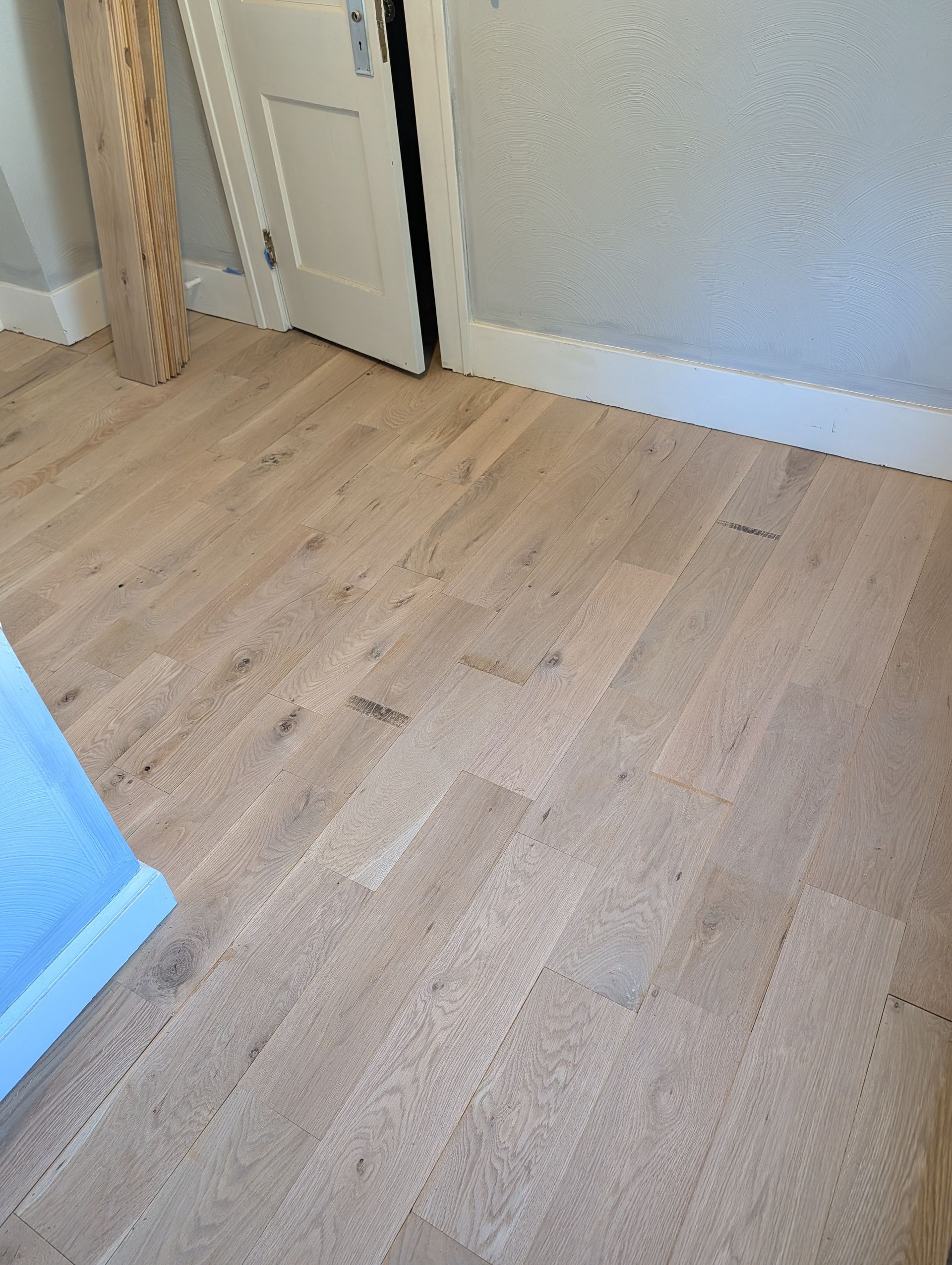 Wooden bedroom floor beside a white door and light gray wall, with a blue bed edge visible.