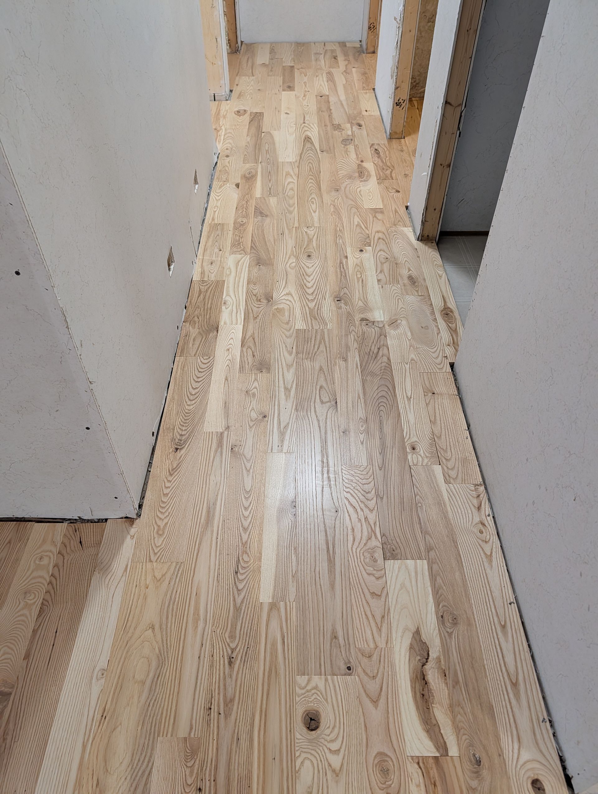Narrow hallway with unfinished wooden floor and white walls leading to a doorway