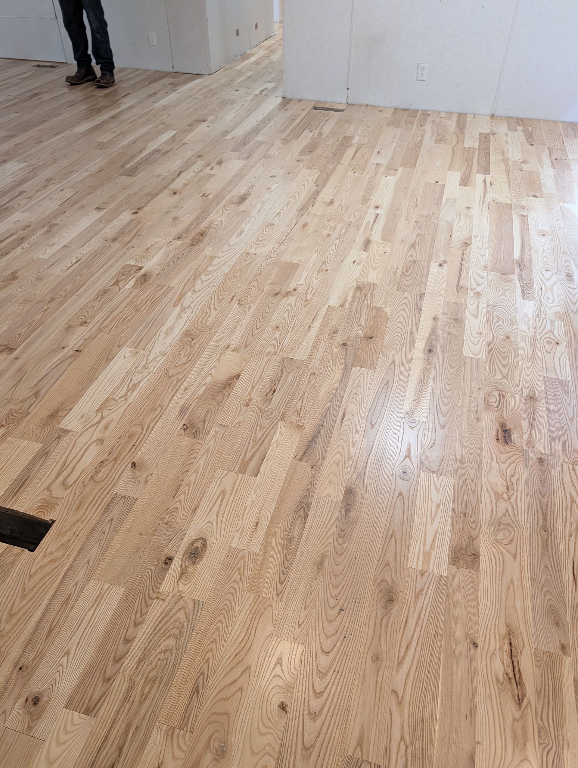 Empty room with light wood flooring and white walls, with a person partially visible at the left edge.