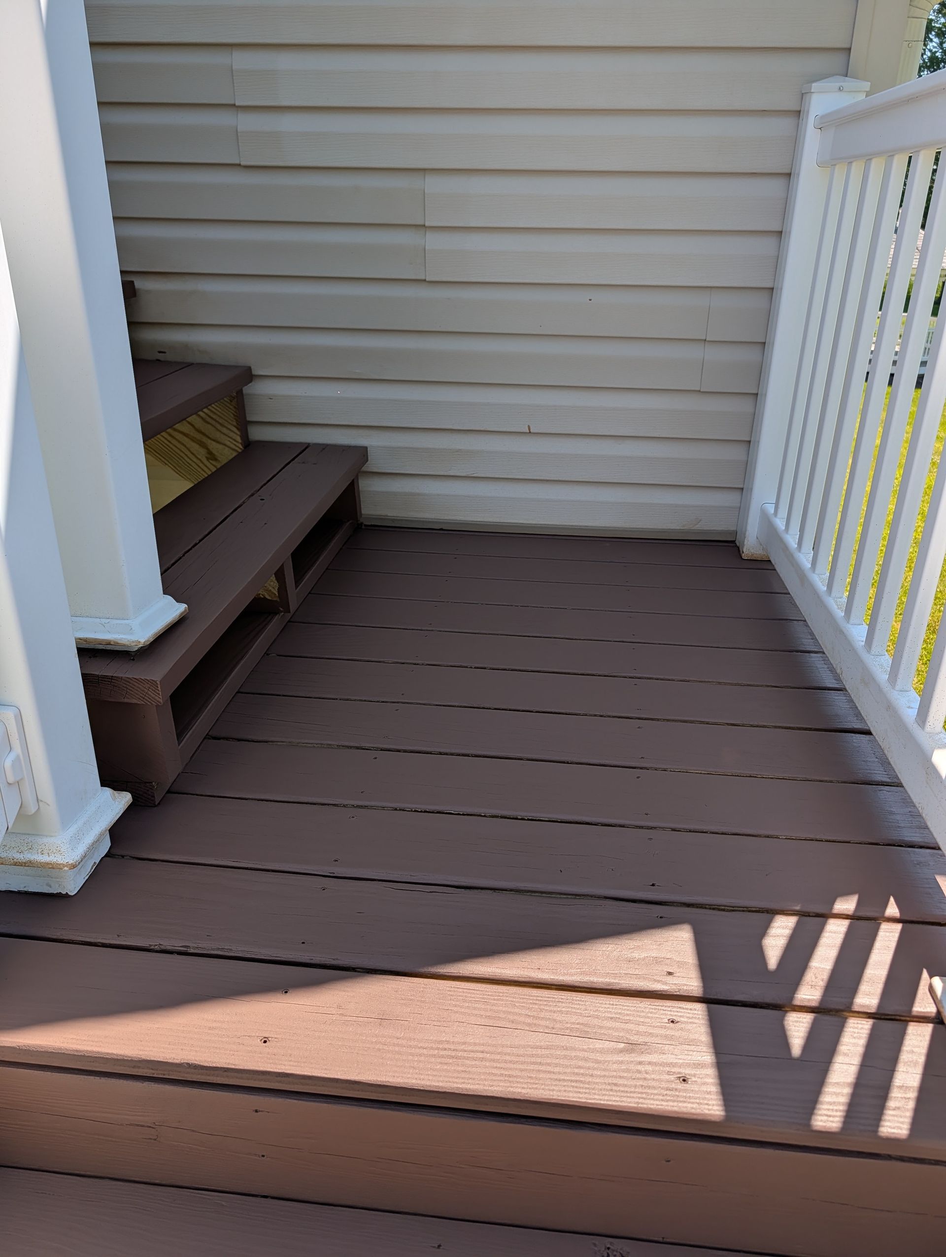 Small brown porch with steps, white railings, and beige siding in bright sunlight.