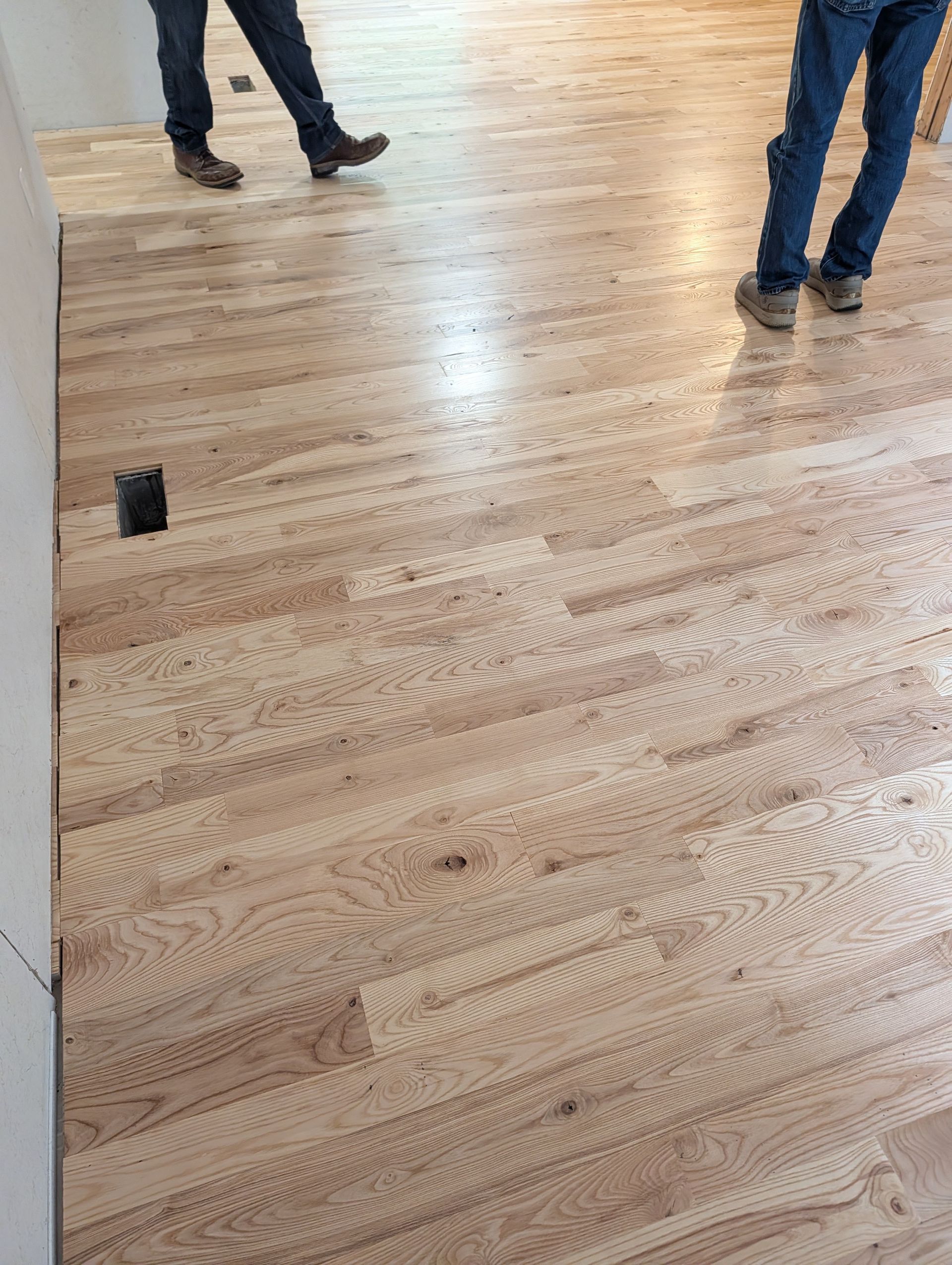 Wooden floor in a room, with two people’s legs and feet visible near the top.