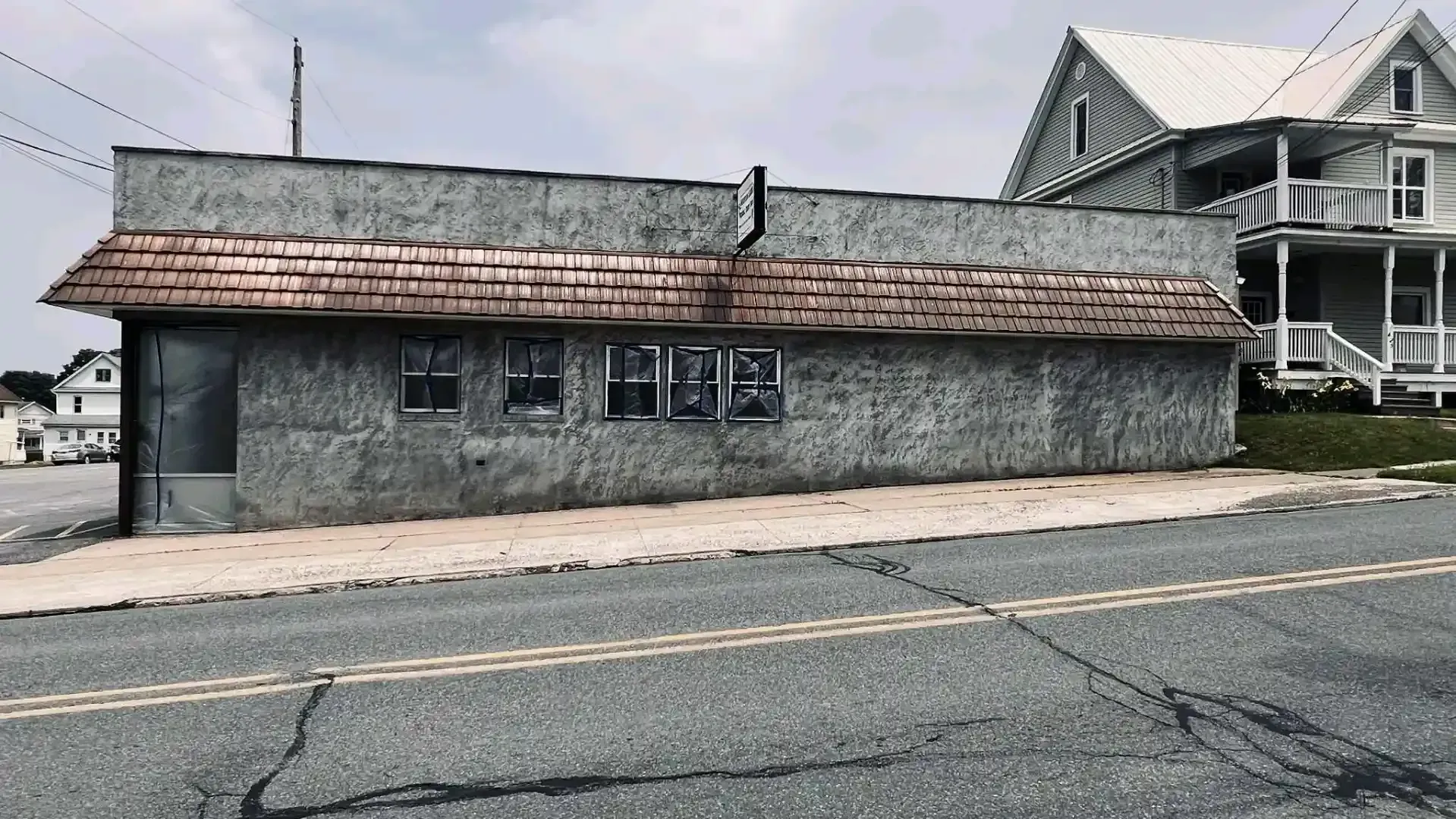 Old gray roadside building with a weathered shingle roof beside a paved street, house in background