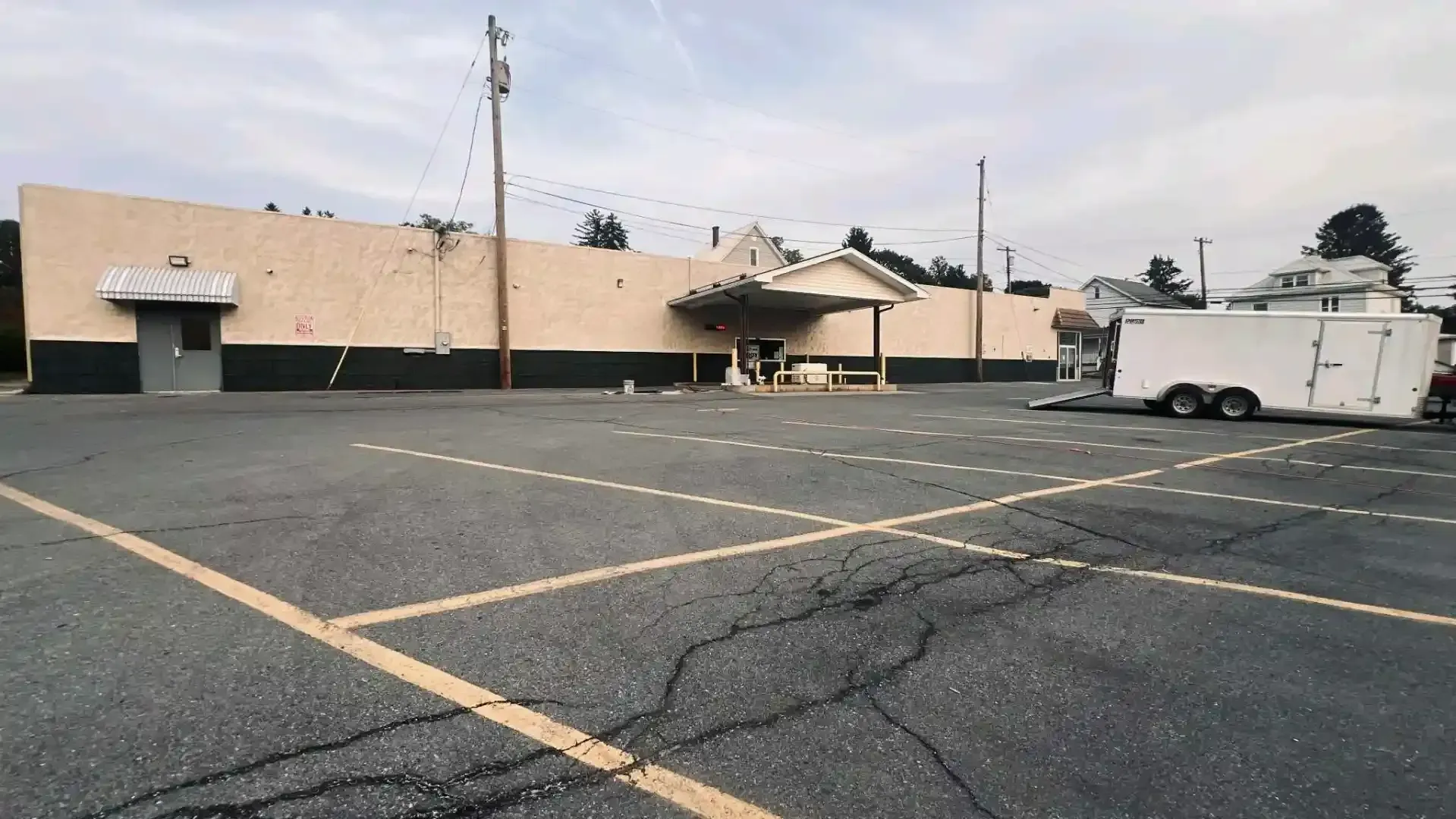 Empty parking lot beside a beige warehouse under an overcast sky, with a white trailer at right.