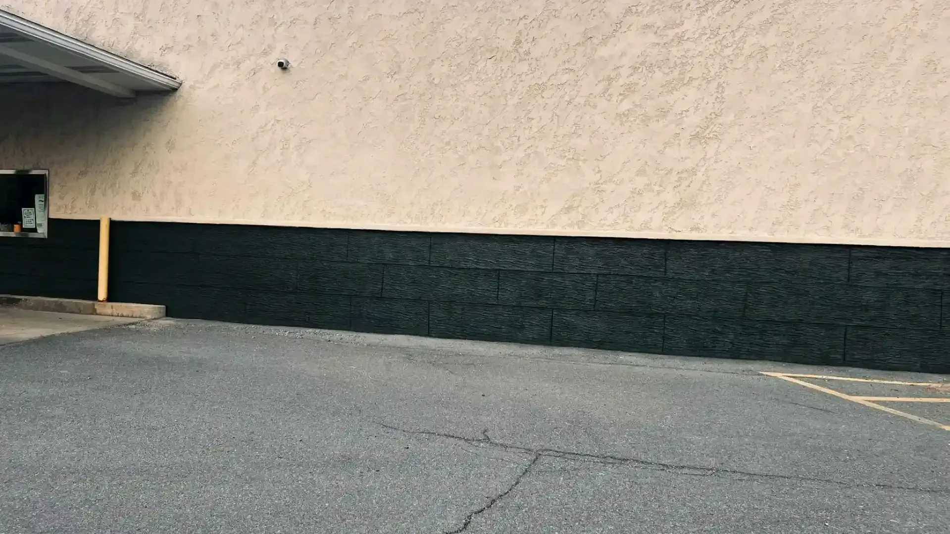 Empty parking lot beside a beige building with a dark lower wall and cracked asphalt pavement