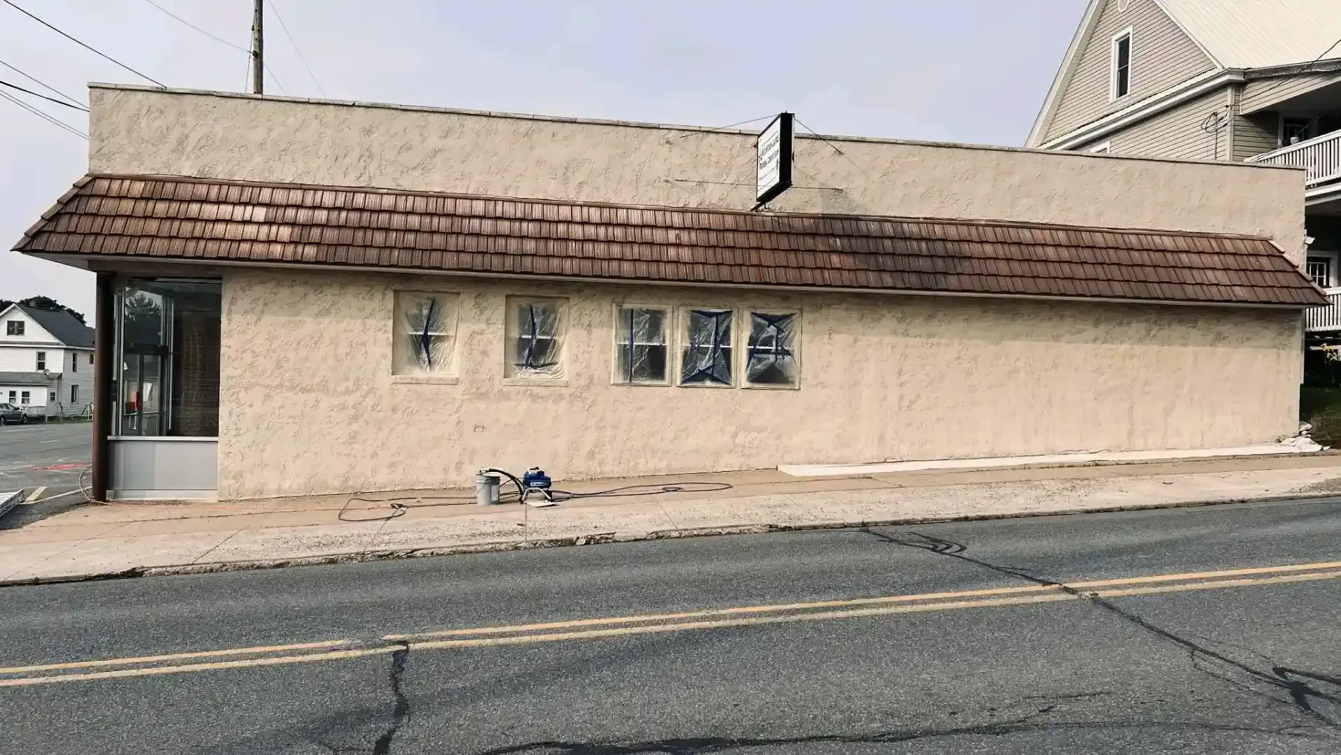 Single-story beige building with a brown shingled roof beside a road, under an overcast sky.