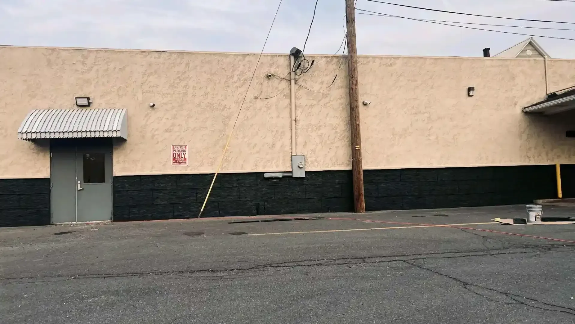 Beige industrial building with a black lower facade, metal door, and empty paved lot in front.