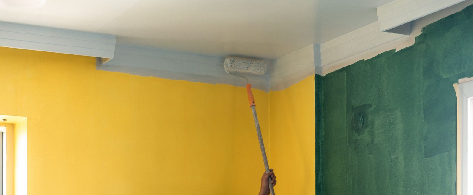 Person painting a room corner with yellow and green walls, using a roller near the ceiling.