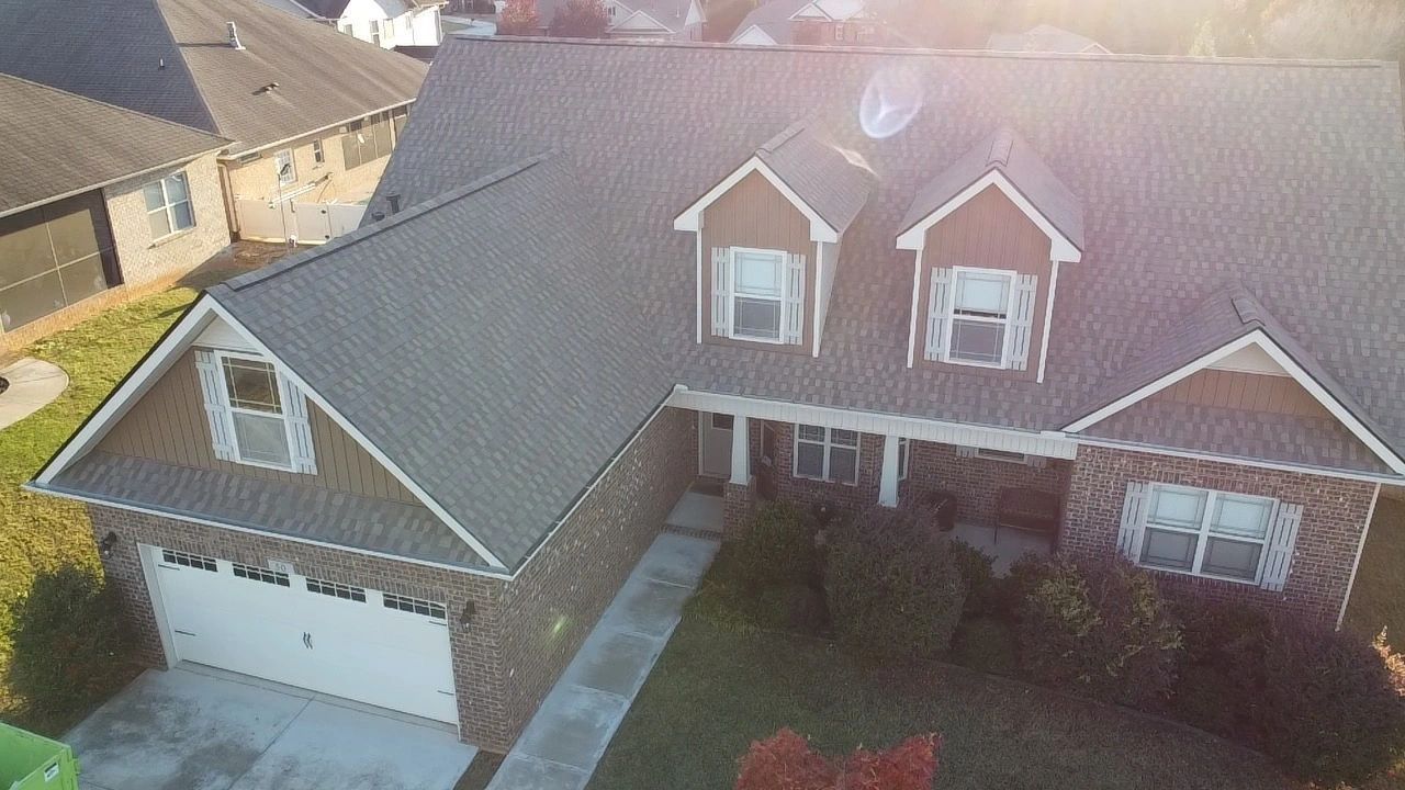 Aerial view of a suburban brick house with a two-car garage, front porch, and gray shingled roof.