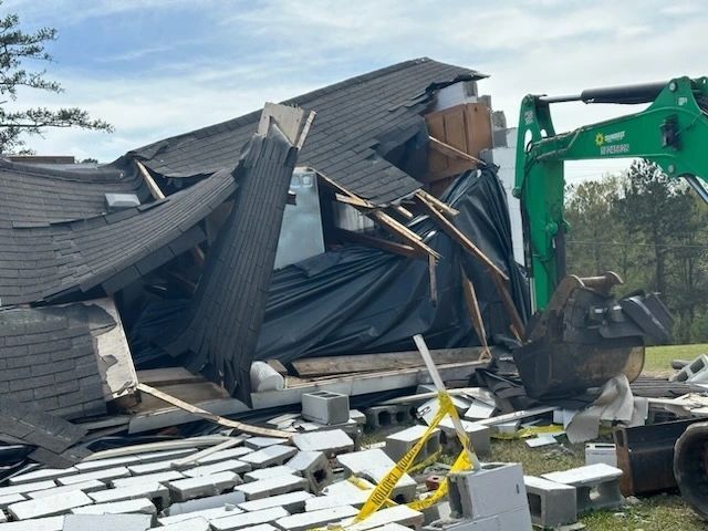 Collapsed house under demolition, with an excavator clearing debris and broken roof shingles scattered in front.