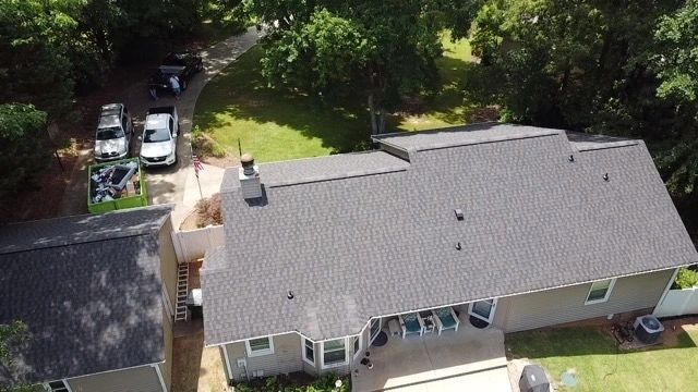 Aerial view of a suburban house with a gray roof, driveway, and trees surrounding the yard