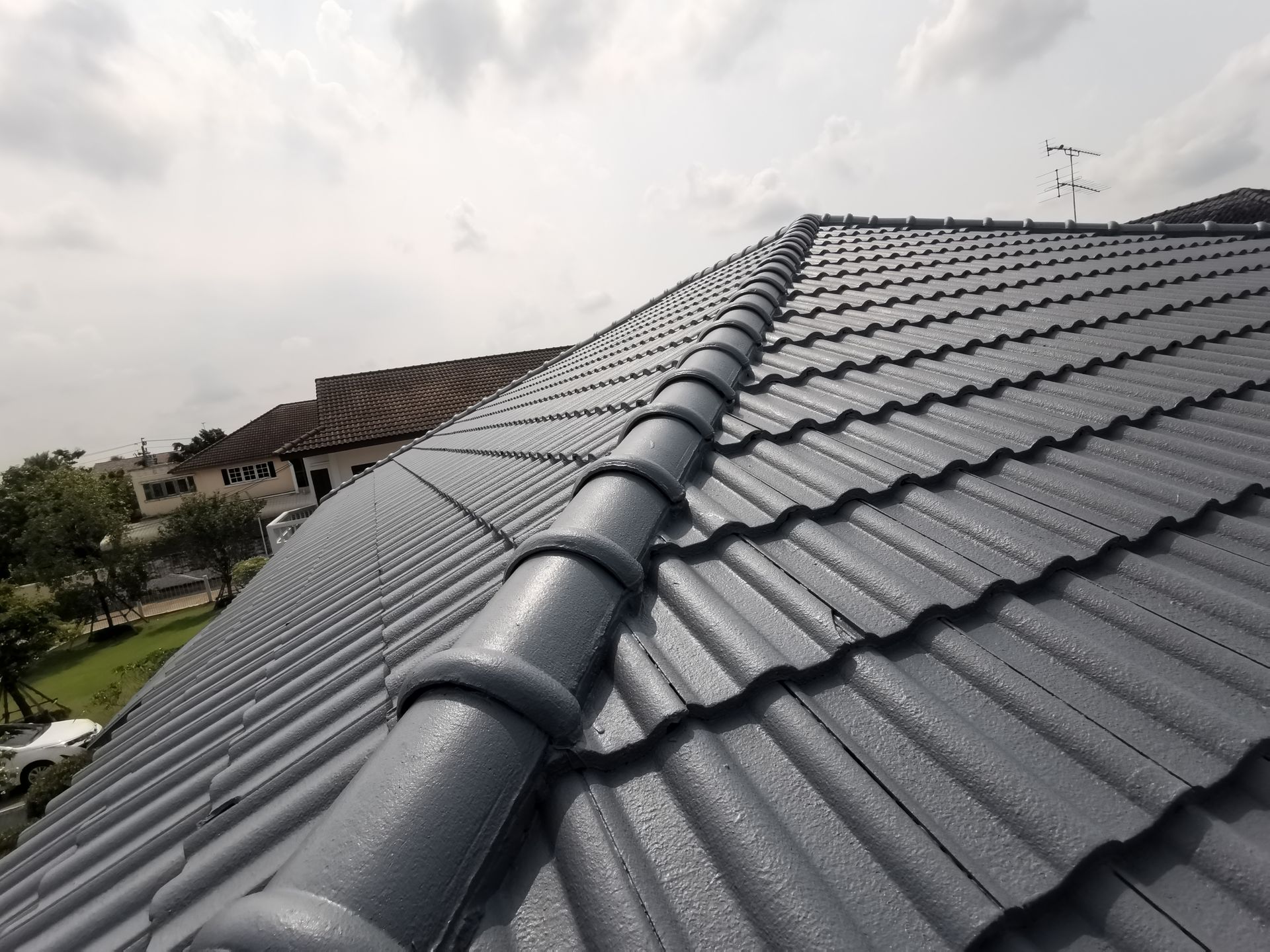 Gray tiled roof with ridge caps viewed from above, with houses and cloudy sky in the background