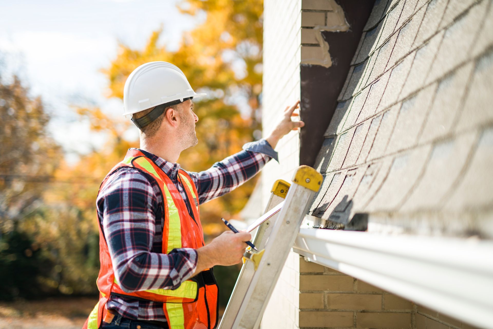 Worker in hard hat and safety vest inspecting house roof with clipboard and hammer