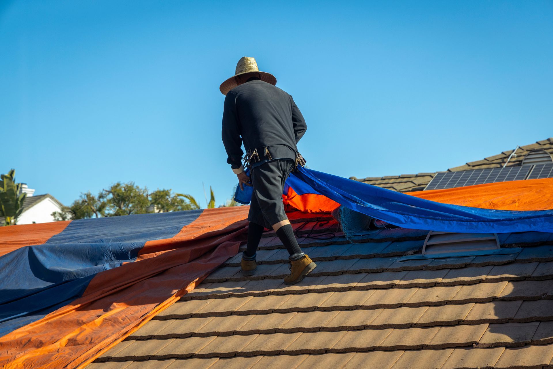Worker spreading orange and blue tarp over a shingled roof under a clear sky