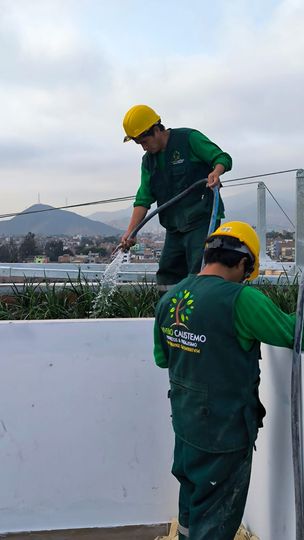 Dos trabajadores con uniformes verdes y cascos amarillos limpian una pared blanca al aire libre