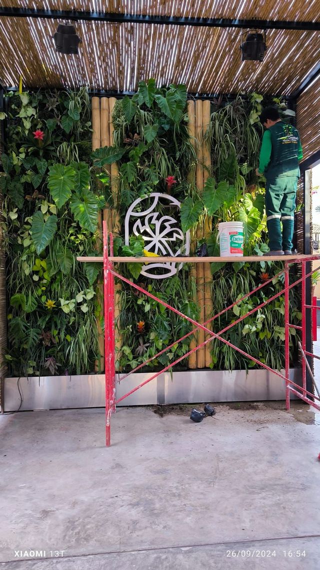 Pared de vegetación exterior con un andamio rojo y adornos circulares colgantes bajo un techo sombreado