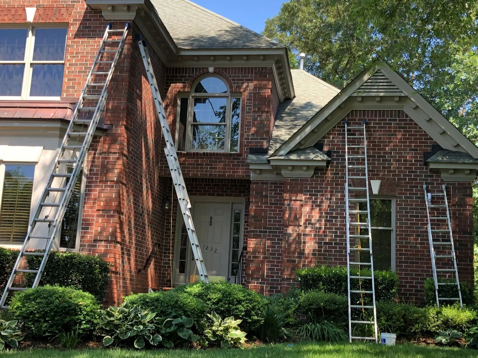 Three ladders leaning against a red brick house; workers are painting the exterior on a sunny day.